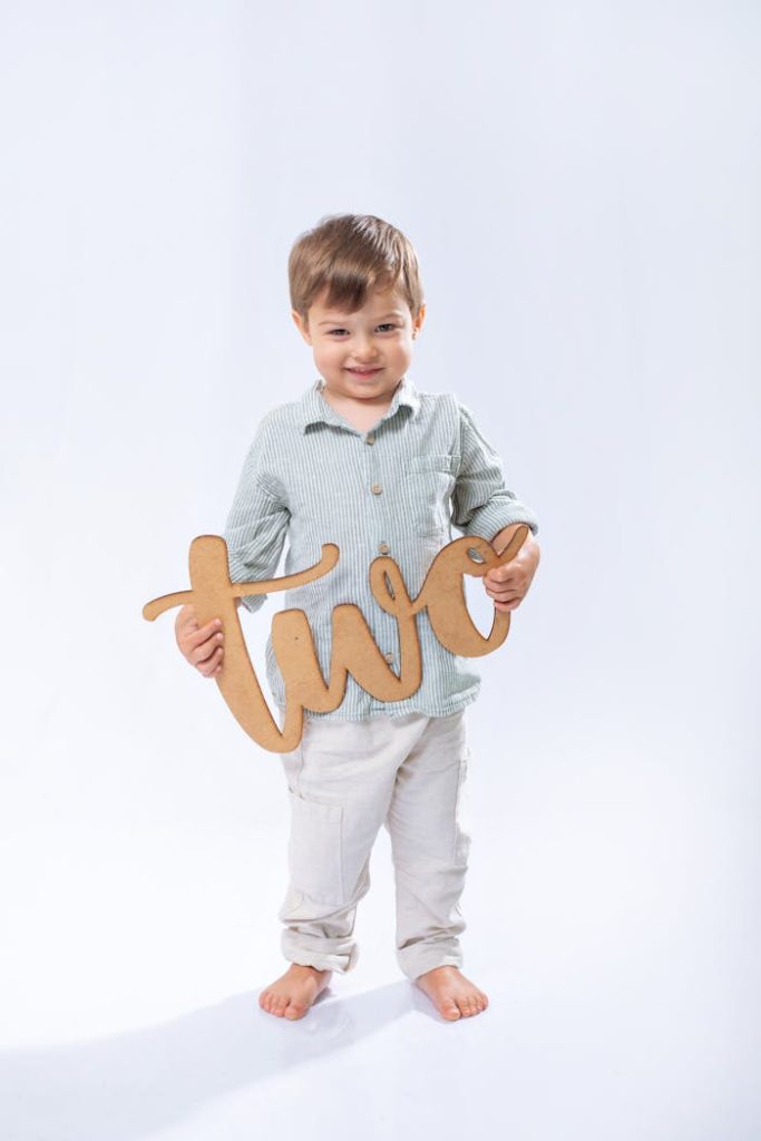 Adorable toddler holding a wooden 'two' sign, smiling on a bright background.