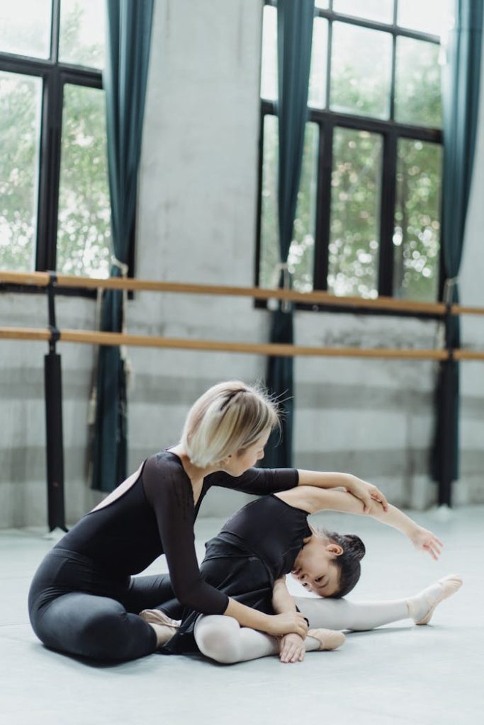 A ballet instructor guides a young girl in stretching during a dance class.