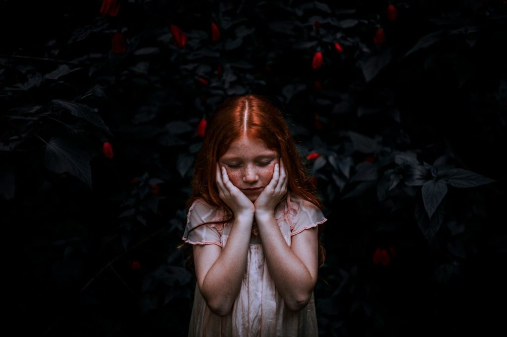 A young redhead girl in a thoughtful pose surrounded by dark foliage.